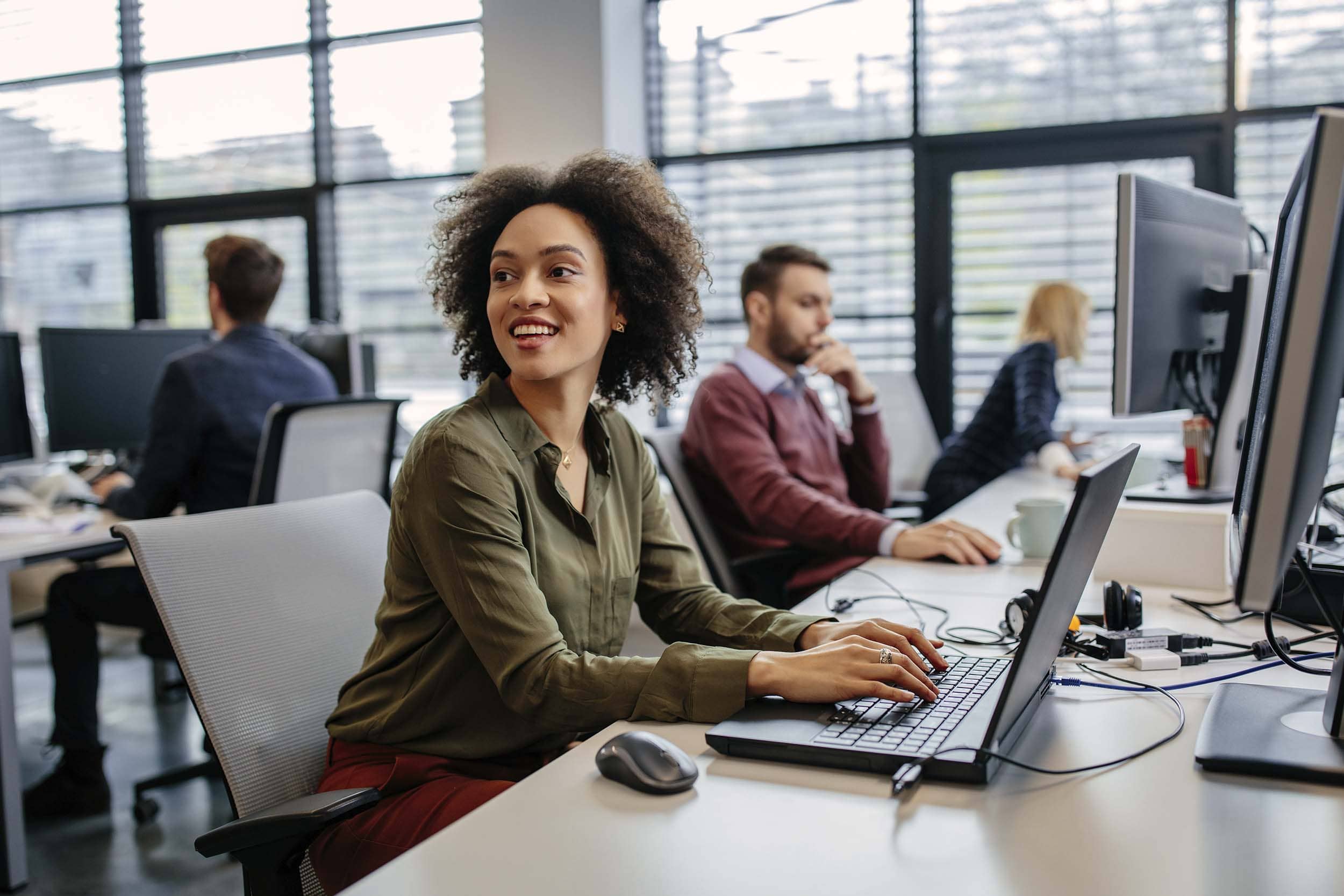 Professional woman smiling while working on a laptop in a busy open-plan hybrid office, reflecting productivity, flexibility, and digital collaboration in a modern workplace.