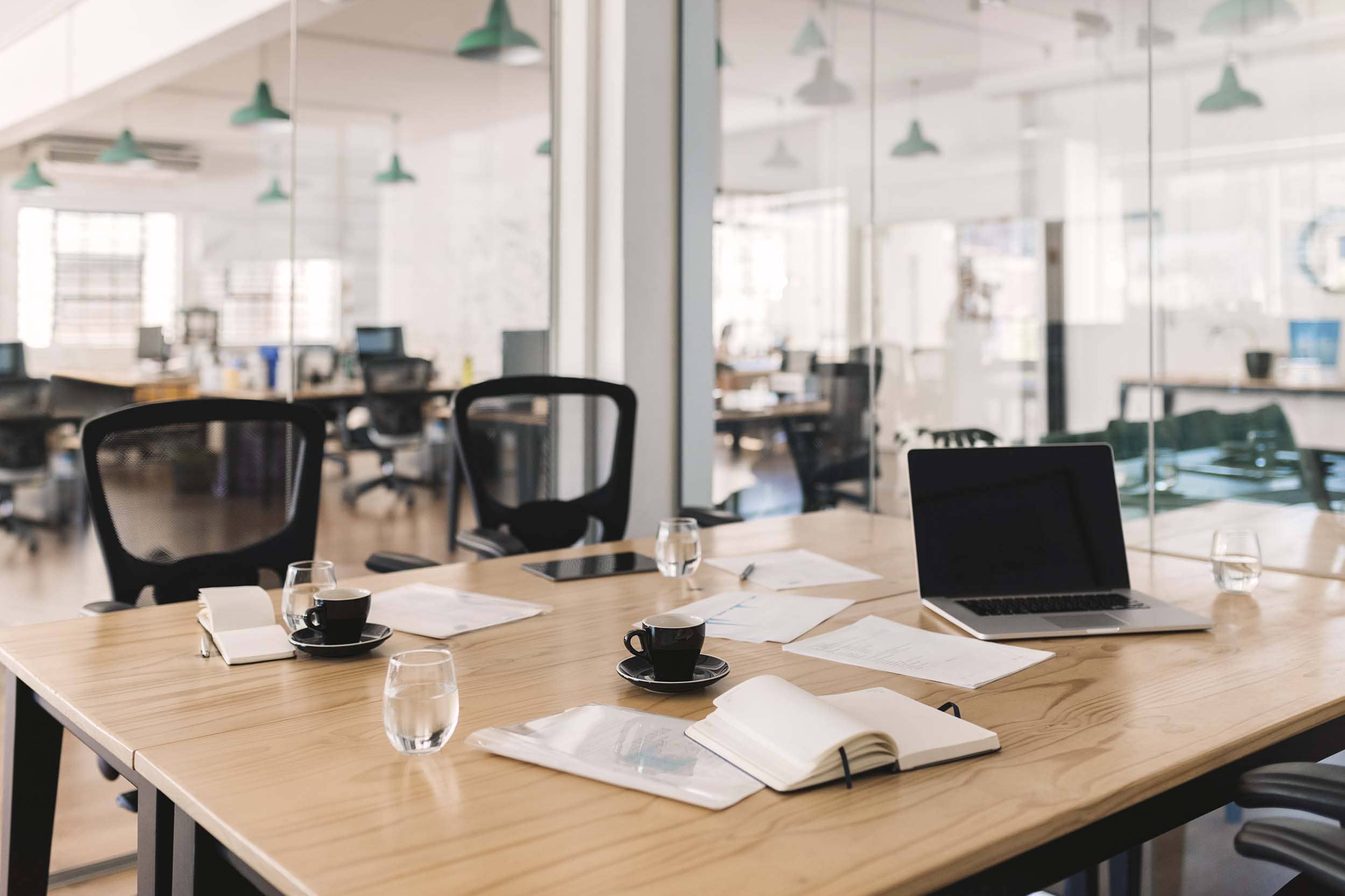 Modern meeting table with open notebooks, coffee cups, and a laptop in a glass-walled hybrid office, symbolizing flexible collaboration and shared workspaces.