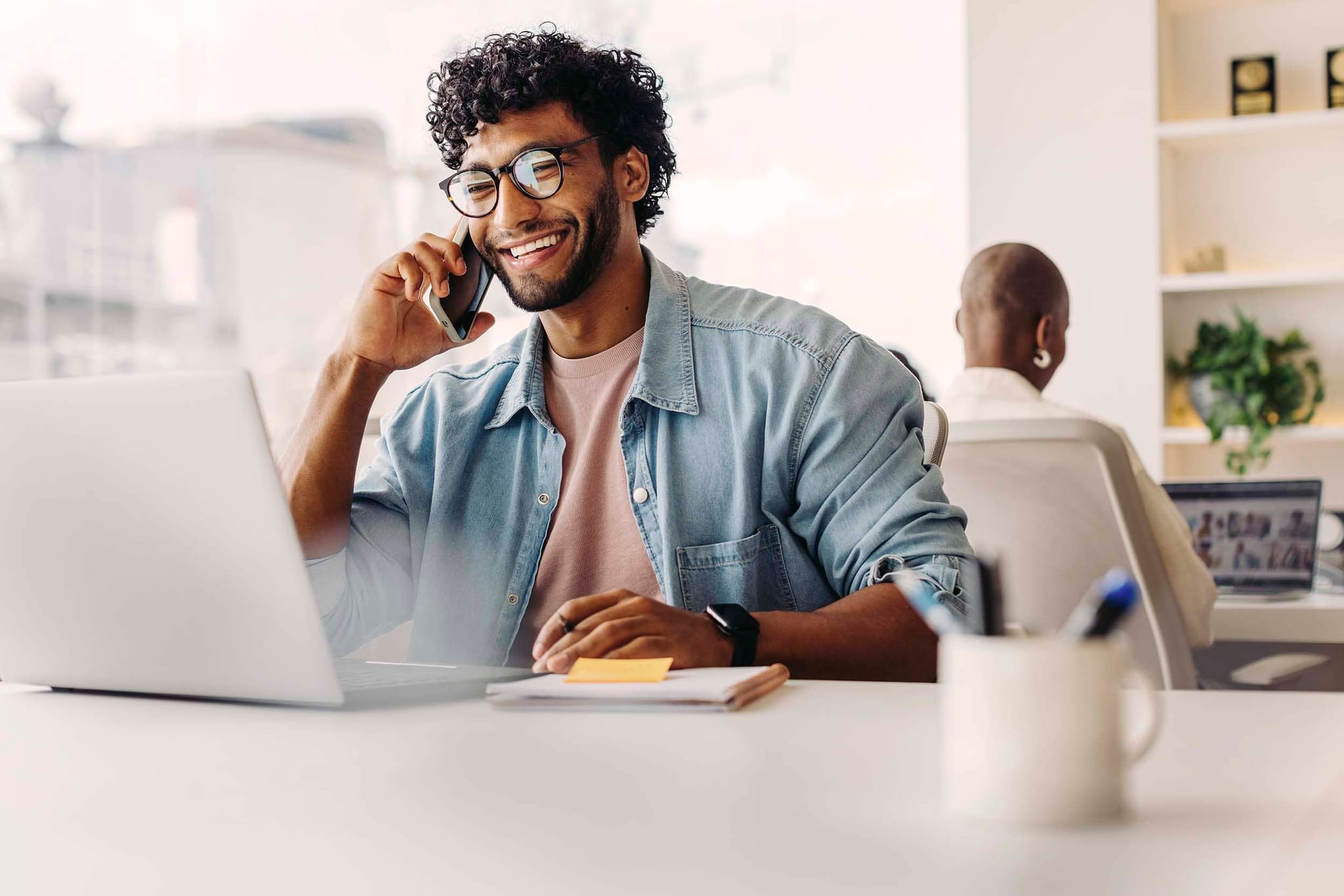 Smiling professional working on a laptop and speaking on the phone in a bright hybrid office setting, representing flexibility and real-time communication in a modern workplace.