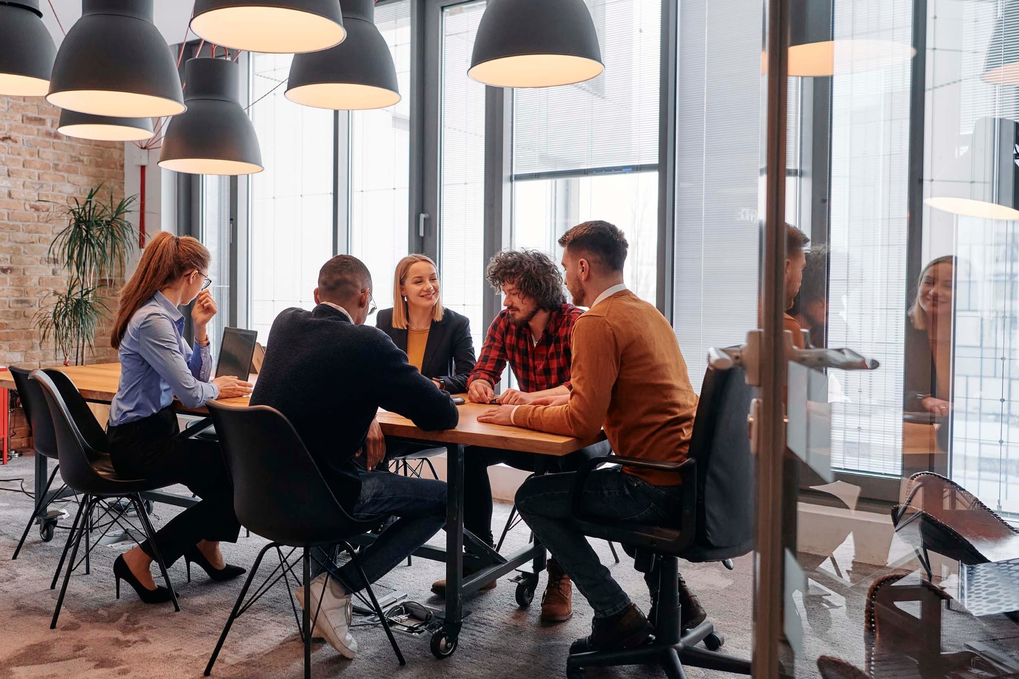 Team of professionals engaged in a meeting around a conference table in a modern glass-walled hybrid office, highlighting collaboration and space utilization in flexible workplaces.