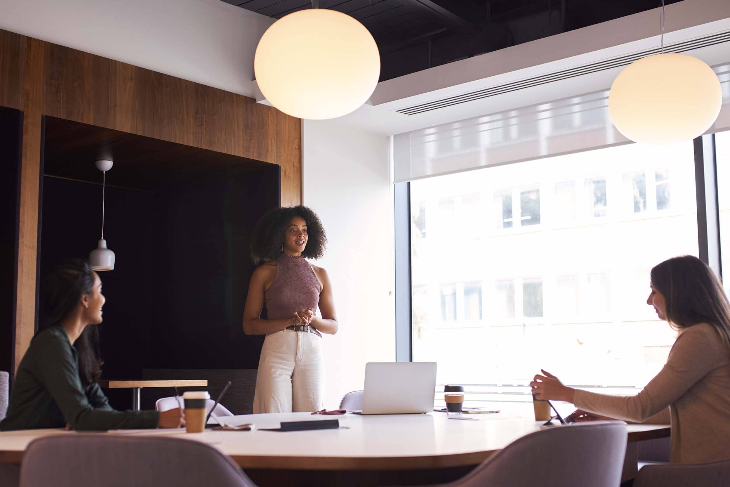 Professional woman leading a meeting in a modern hybrid office conference room, with colleagues seated around a table using laptops and coffee cups, showcasing flexible team collaboration.