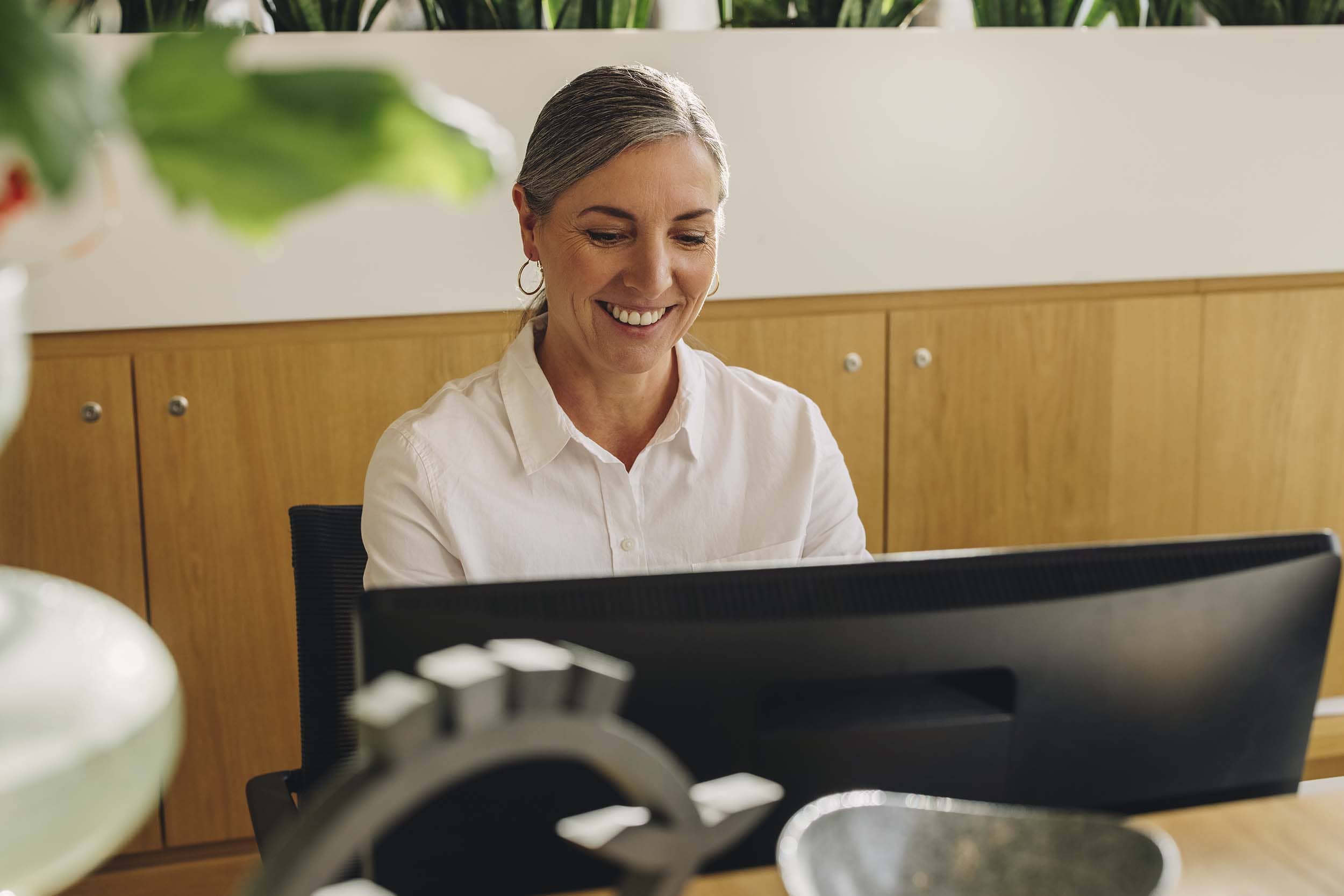 Smiling woman working at a desktop computer in a stylish, plant-filled hybrid office space, reflecting comfort, focus, and productivity in a flexible work environment.
