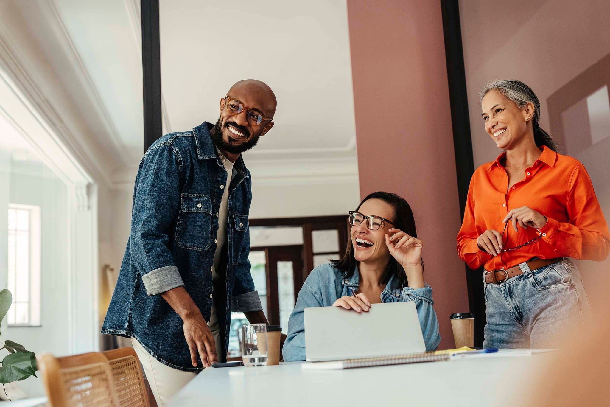 Group of professionals sharing a light moment during a hybrid office meeting, with laptops and coffee on the table, illustrating teamwork and flexibility in a modern work environment.